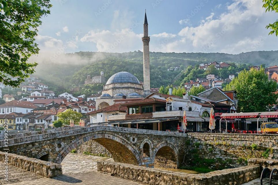 Un puente de arco de piedra cruza un río frente a un pueblo con una mezquita y un minarete, con una colina verde y brumosa al fondo.