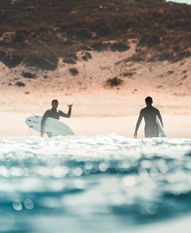 Un viaggio di gruppo WeRoad di due surfisti in muta con le loro tavole nell'oceano, uno che fa il segno shaka.
