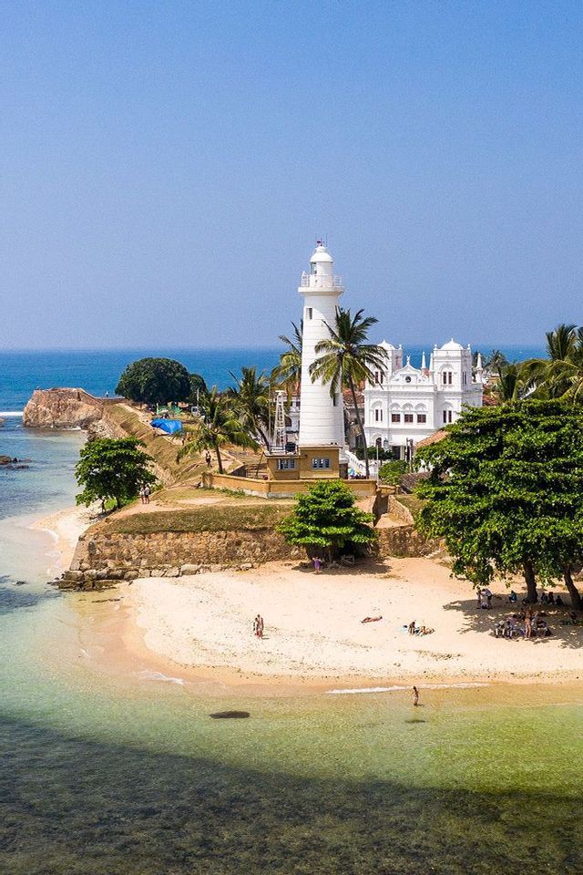 Un faro bianco si erge su una costa fortificata sopra una spiaggia sabbiosa con palme e acqua cristallina.