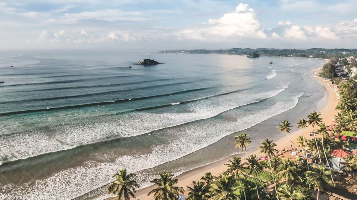 Veduta aerea di una costa tropicale con spiaggia di sabbia, palme e surfisti su onde dolci.