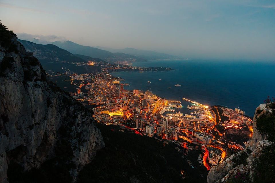 Veduta dall'alto di una città costiera illuminata e del suo porto turistico al tramonto, incastonata tra mare e monti.