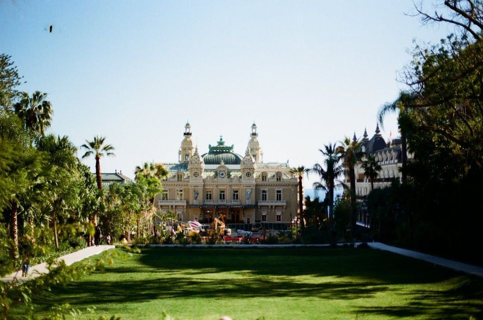 Una vista del Casinò di Monte Carlo da un parco, con un grande prato verde in primo piano e palme che incorniciano l'edificio.