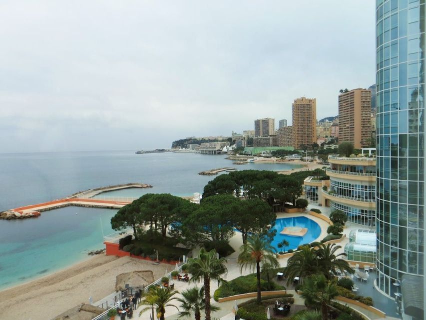 Una vista dall'alto di una città costiera con una spiaggia sabbiosa, una grande piscina e edifici moderni sotto un cielo coperto.