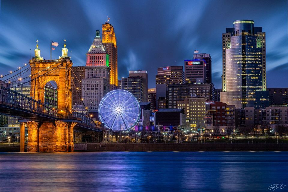 Un ponte illuminato e una ruota panoramica sullo skyline di una città di notte, visti da oltre uno specchio d'acqua.