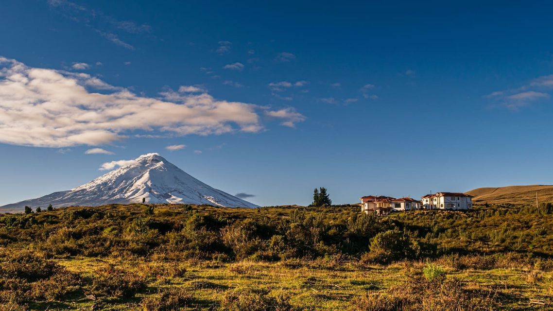 Un vulcano innevato con un pennacchio di fumo che si alza dalla cima, situato dietro una collina erbosa con diverse case bianche sotto un cielo azzurro.