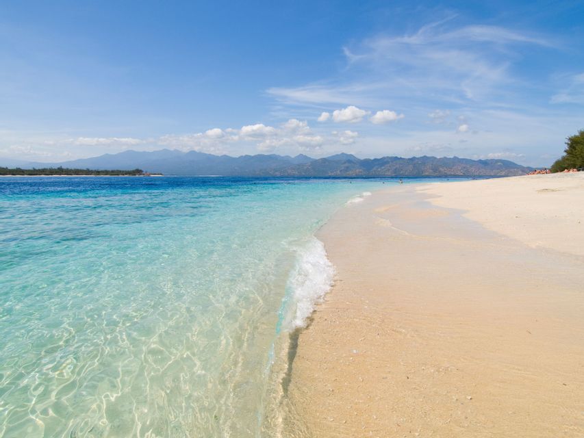 Une eau turquoise claire vient doucement lécher une plage de sable blanc avec des montagnes en arrière-plan sous un ciel bleu.