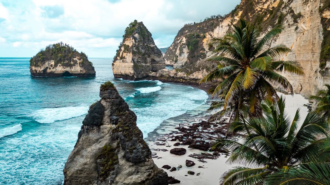 A view of a tropical cove with turquoise water, a white sand beach, and large rock formations topped with greenery.