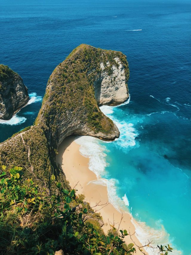 An aerial view of a unique T-Rex shaped cliff covered in green foliage, overlooking a secluded white sand beach and turquoise sea.