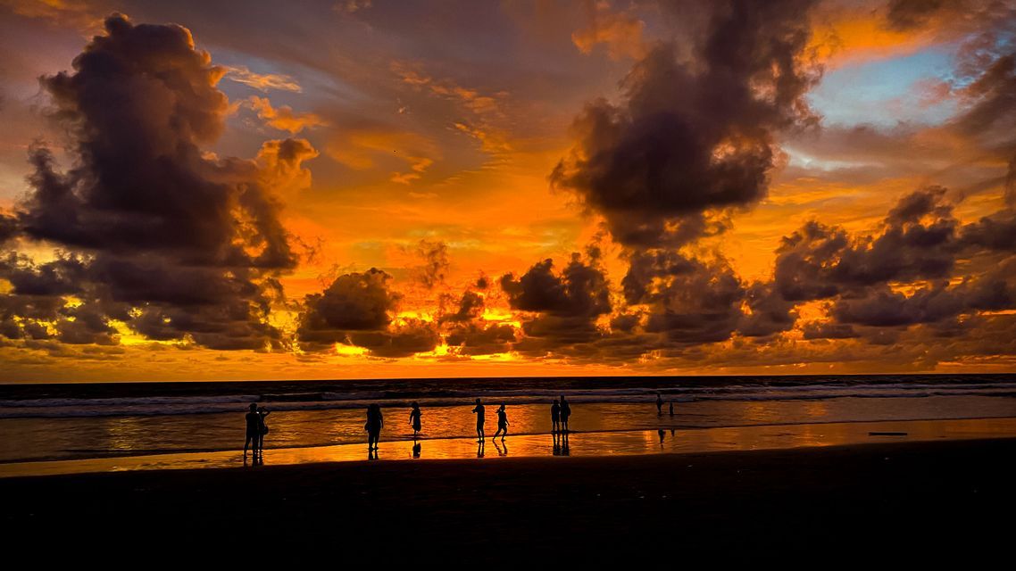 Un groupe de voyage WeRoad se découpant sur une plage mouillée, regardant un coucher de soleil orange spectaculaire avec des nuages sombres sur l'océan.