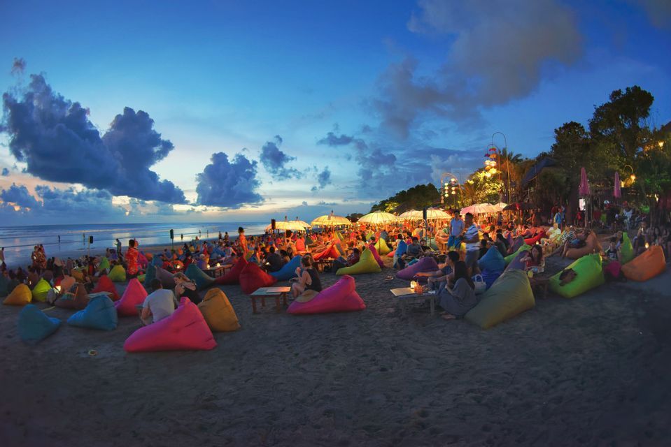 Un groupe de WeRoad se détend sur des poufs colorés sur une plage de sable au crépuscule, avec des parasols illuminés au-dessus.