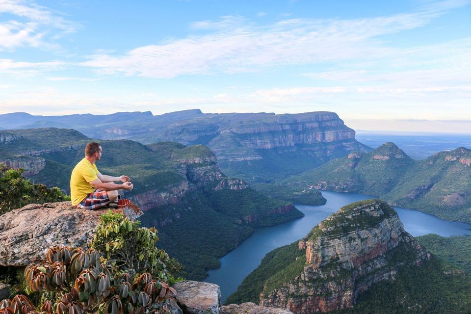 Un uomo con una maglietta gialla siede a gambe incrociate su una scogliera rocciosa, contemplando un vasto canyon verde e un fiume sinuoso che scorre sotto.