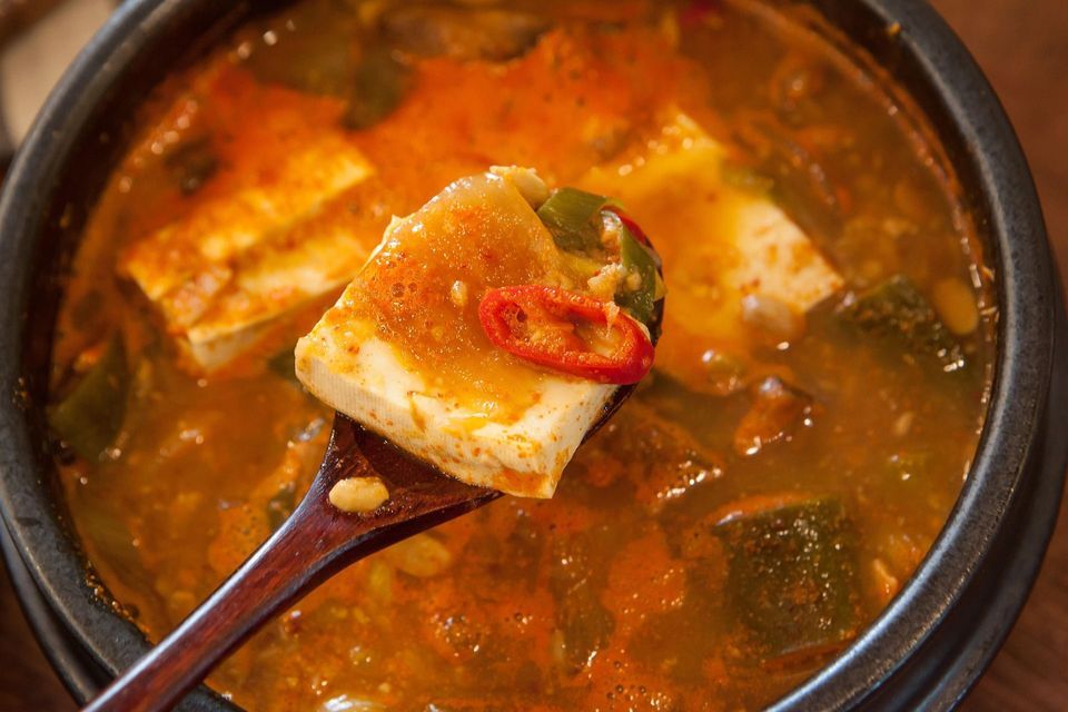 Un cucchiaio di legno solleva un pezzo di tofu con una fetta di peperoncino rosso da uno stufato arancione bollente in una ciotola.