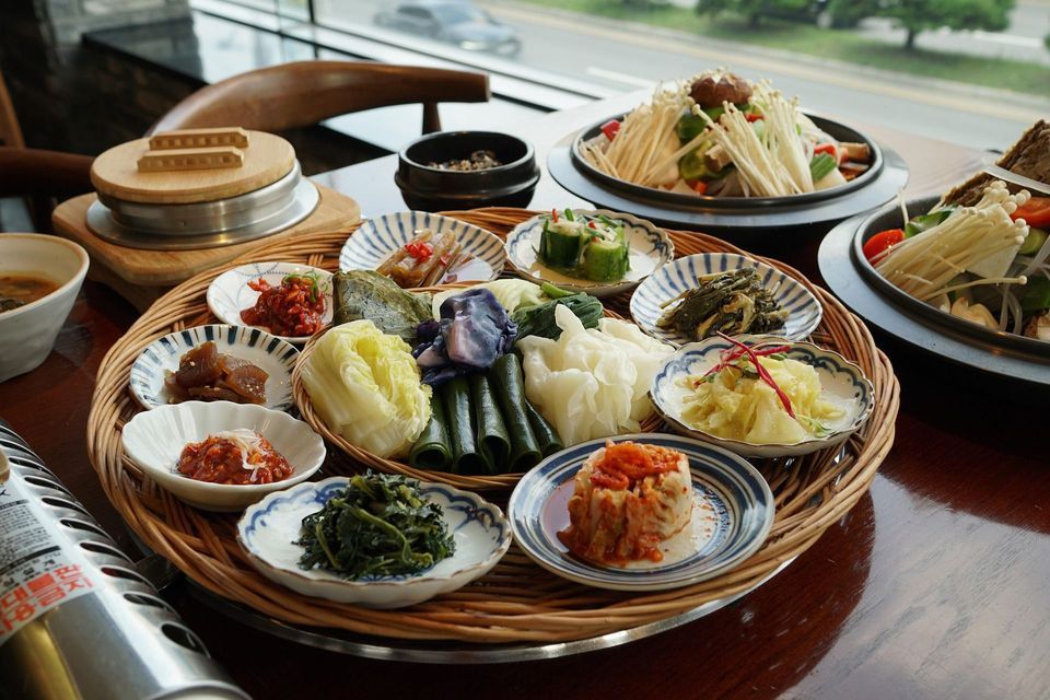 Un tradizionale pasto coreano con vari contorni (banchan) in un cesto intrecciato e un hot pot di funghi su un tavolo di legno.