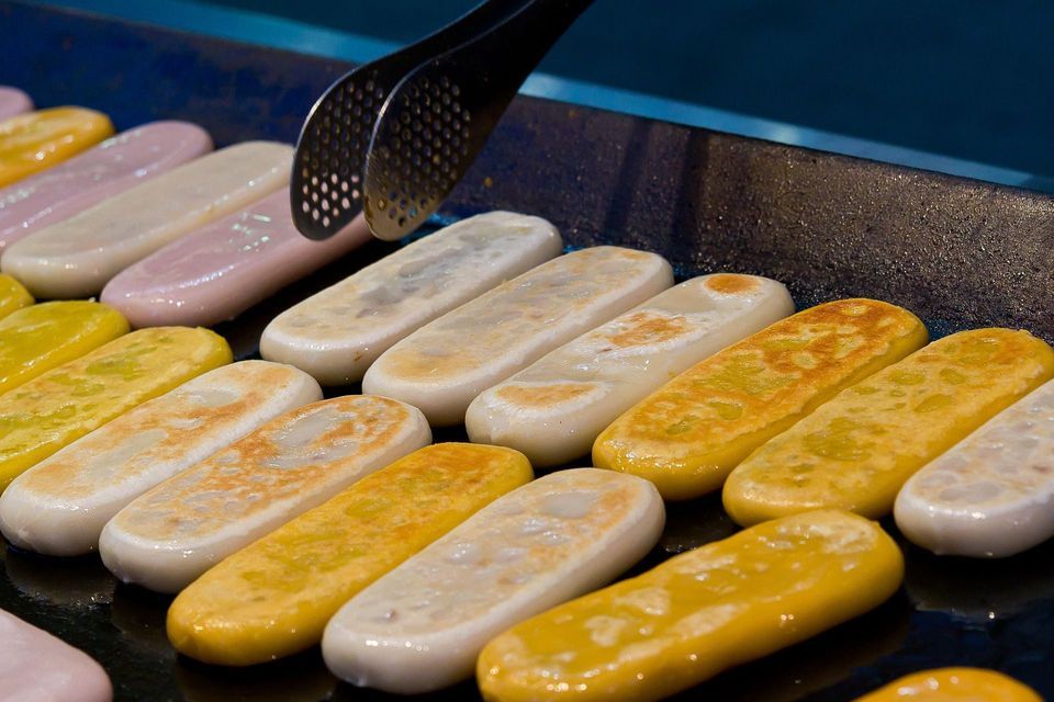 Primo piano di gnocchi di riso oblunghi e colorati, fritti in padella in file ordinate su una piastra scura, con pinze metalliche sopra.