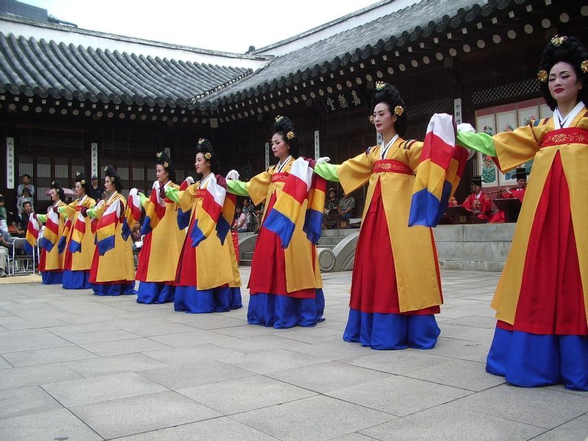 Una fila di donne in costumi tradizionali coreani colorati esegue una danza in un cortile storico.