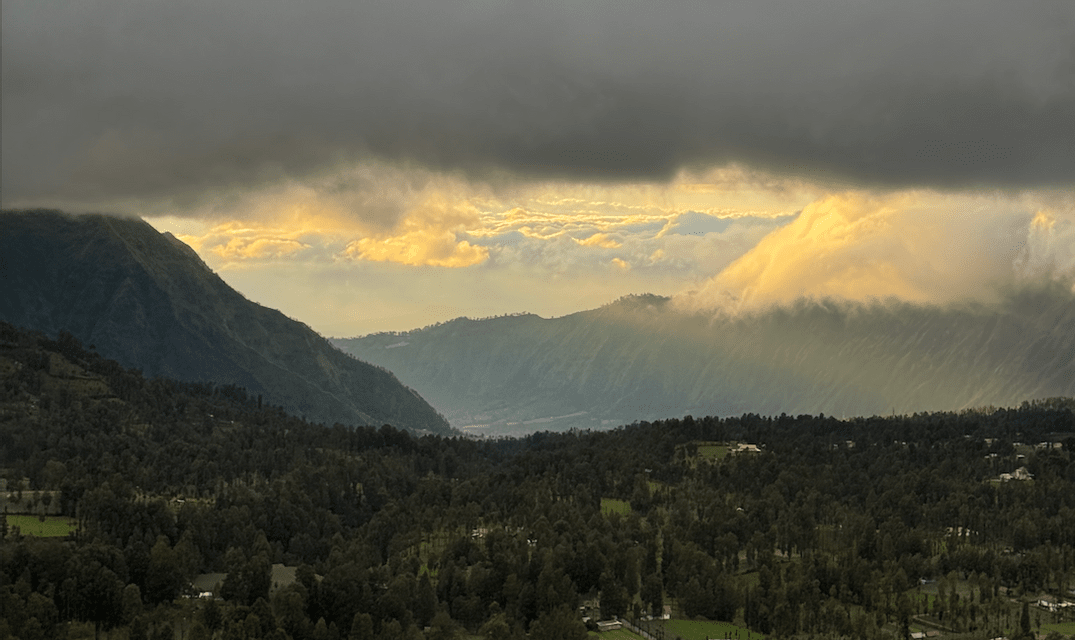 Ein üppiges grünes Tal und bewaldete Berge unter einem dunklen, bewölkten Himmel mit durchbrechendem goldenem Sonnenlicht.