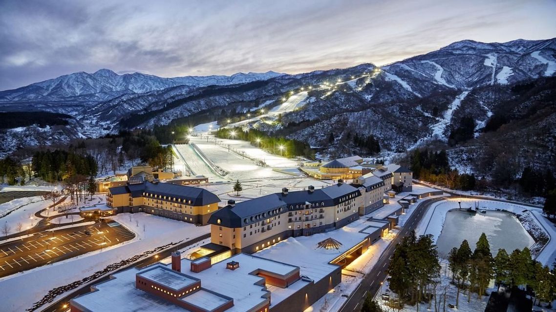 Luftaufnahme eines großen, beleuchteten Skigebiets am Fuße schneebedeckter Berge in der Dämmerung.