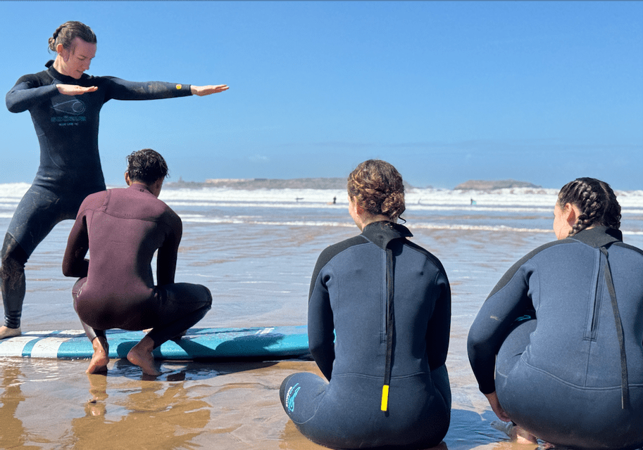 Ein Surflehrer im Neoprenanzug demonstriert einer WeRoad-Gruppe an einem sonnigen Strand die Haltung während einer Surfstunde.