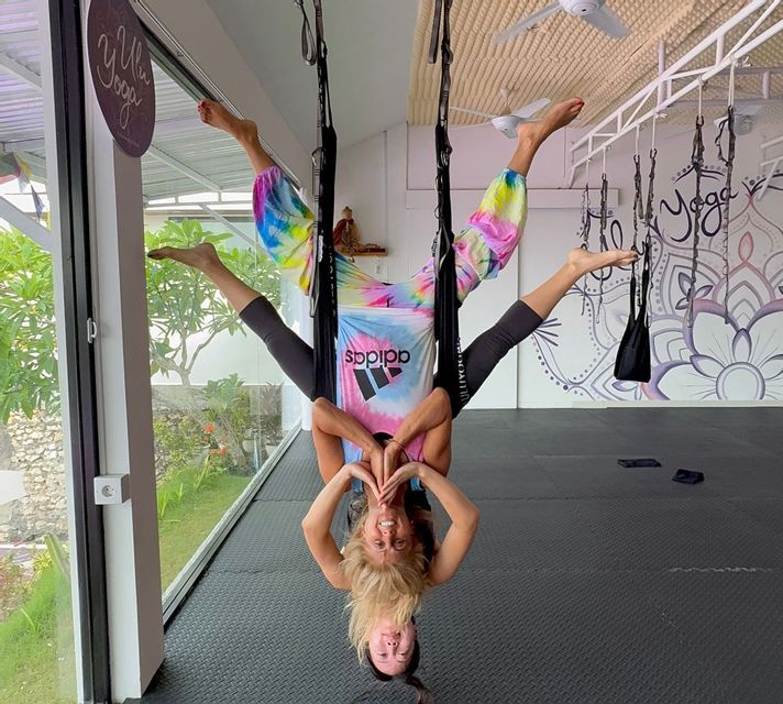 Zwei Frauen hängen kopfüber in schwarzen Aerial-Yoga-Hängematten in einem Studio und lächeln in die Kamera.