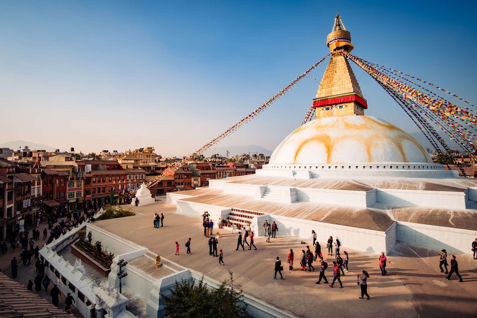 Ein Blick von oben auf Menschen, die auf den Terrassen einer großen, weißen buddhistischen Stupa mit einer goldenen Spitze und farbenfrohen Gebetsfahnen gehen.