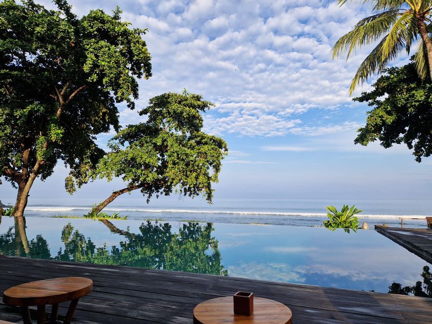 Ein Infinity-Pool auf einem Holzdeck mit Blick auf den Ozean, der Bäume und einen teilweise bewölkten blauen Himmel spiegelt.