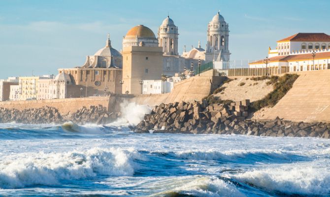 Onde oceaniche si infrangono contro una diga di pietra che protegge una città costiera con una grande cattedrale sotto un cielo limpido.