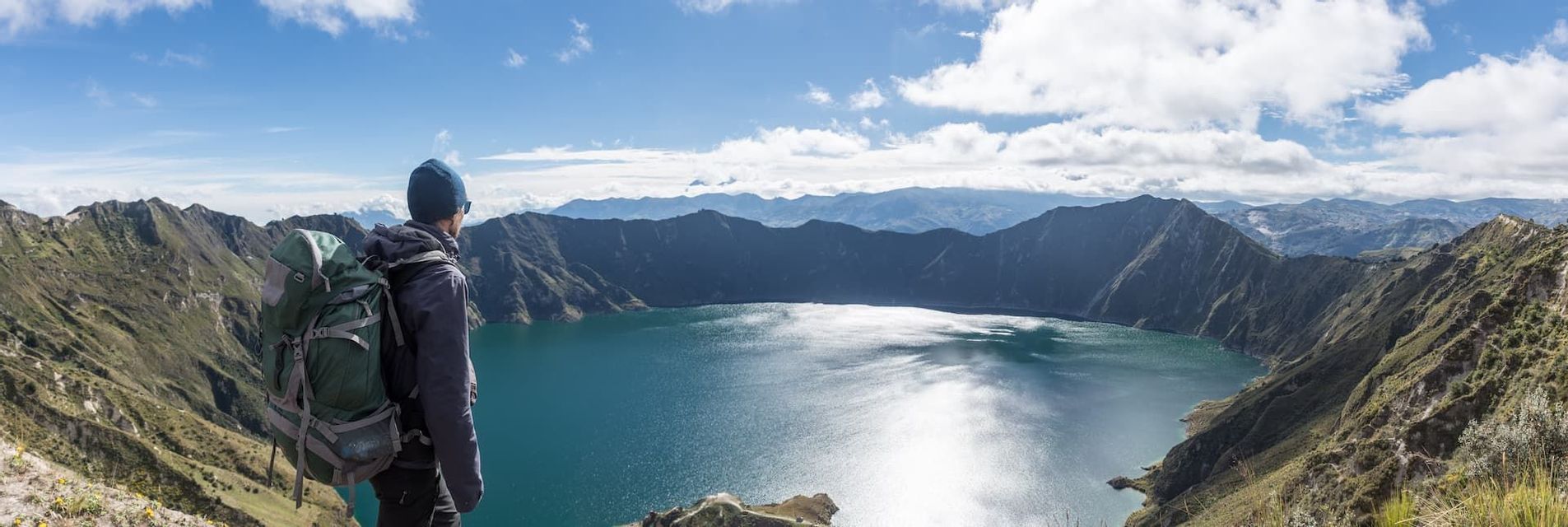 Ein Wanderer mit Rucksack steht auf einem Berggrat und blickt auf einen weiten Kratersee unter einem teils bewölkten blauen Himmel.