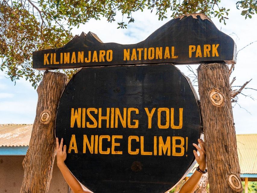 A person's hands hold the wooden sign for Kilimanjaro National Park, which reads, "Wishing you a nice climb!".