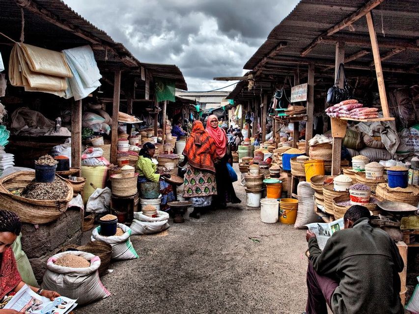 A busy outdoor market alley with people browsing stalls filled with grains and spices in woven baskets.