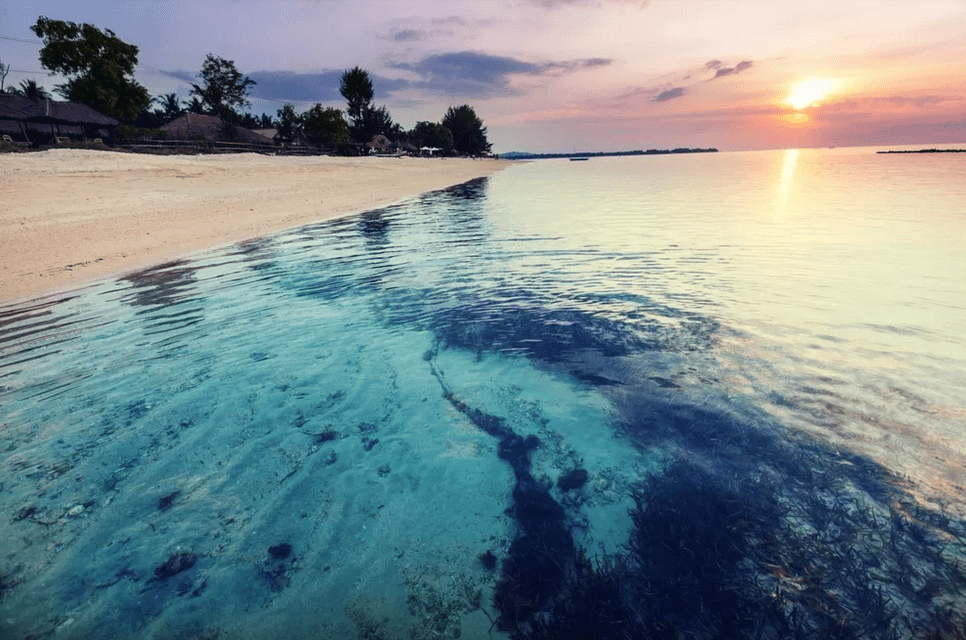 Clear turquoise water with a visible seabed of sand and seaweed laps against a sandy beach at sunset.
