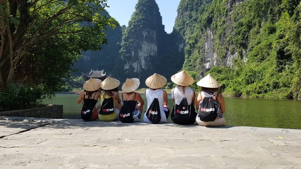Un grupo de viaje WeRoad, sentado en una saliente de piedra, con sombreros cónicos, contempla un río rodeado de exuberantes montañas verdes.