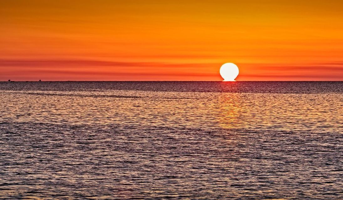 Le soleil se couche sous l'horizon de l'océan, se reflétant sur l'eau sous un ciel vibrant d'orange et de rouge.