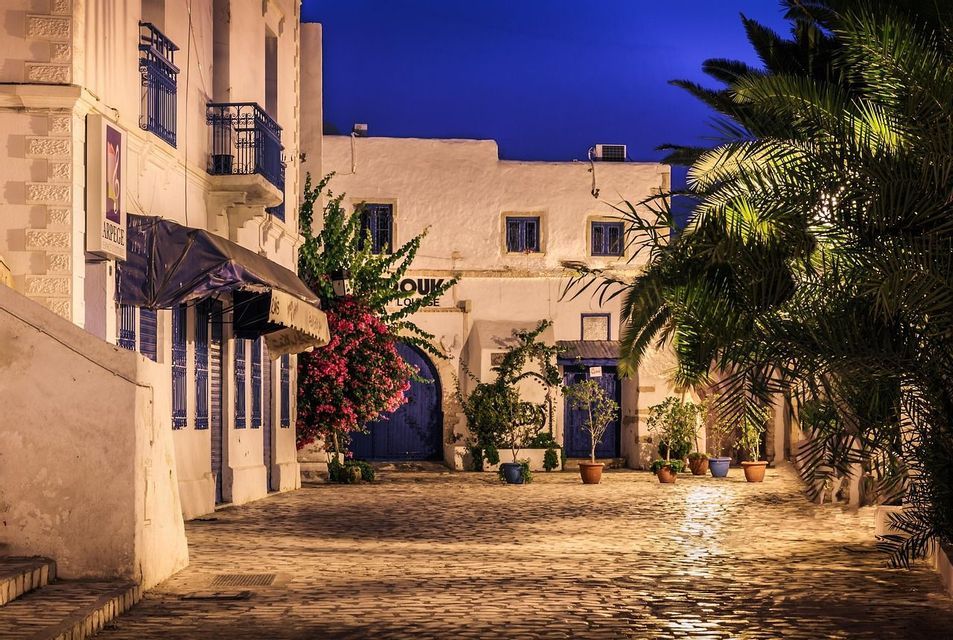 Un patio pavé illuminé par des lumières chaudes la nuit, entouré de bâtiments blancs avec des portes bleues et des palmiers.
