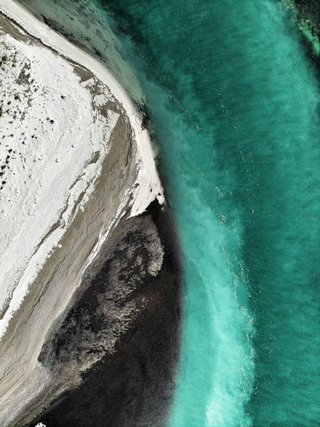 Vue aérienne d'une plage de sable blanc bordant une eau turquoise claire, avec des formations de sable foncé.