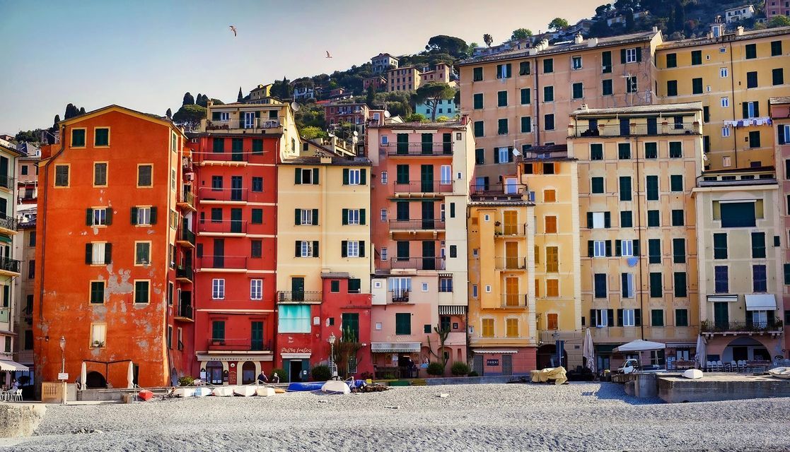 Edifici colorati a più piani costeggiano una spiaggia di ciottoli, con altre case costruite sulla verde collina sullo sfondo.
