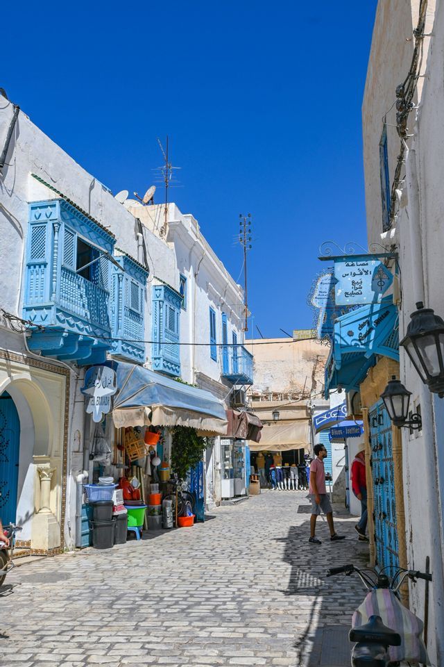Une rue étroite et pavée, baignée de soleil, bordée de bâtiments blancs avec des balcons et des portes bleu vif sous un ciel clair.