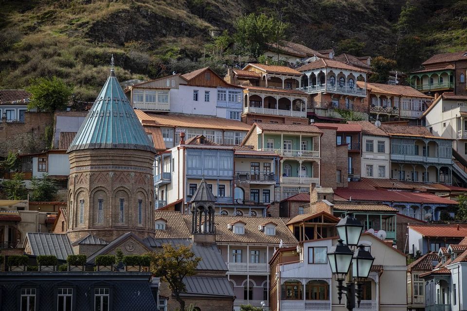 A brick tower with a teal conical roof stands among traditional houses with balconies built on a grassy hillside.