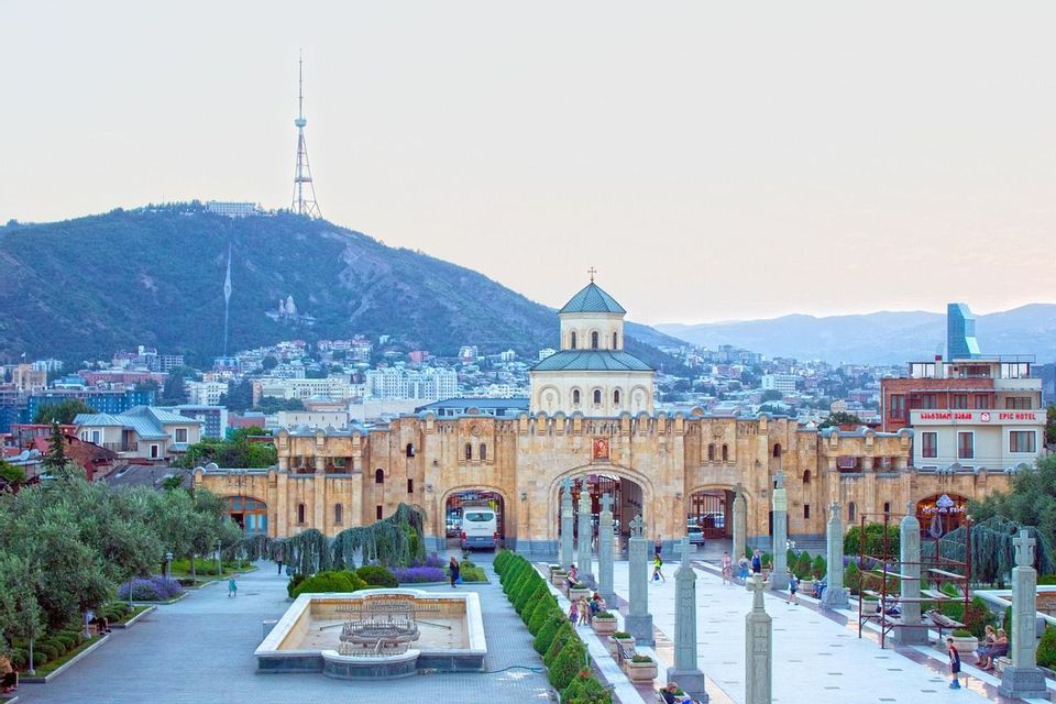 A stone cathedral with a large archway entrance, set against a cityscape with a large hill and a communications tower.