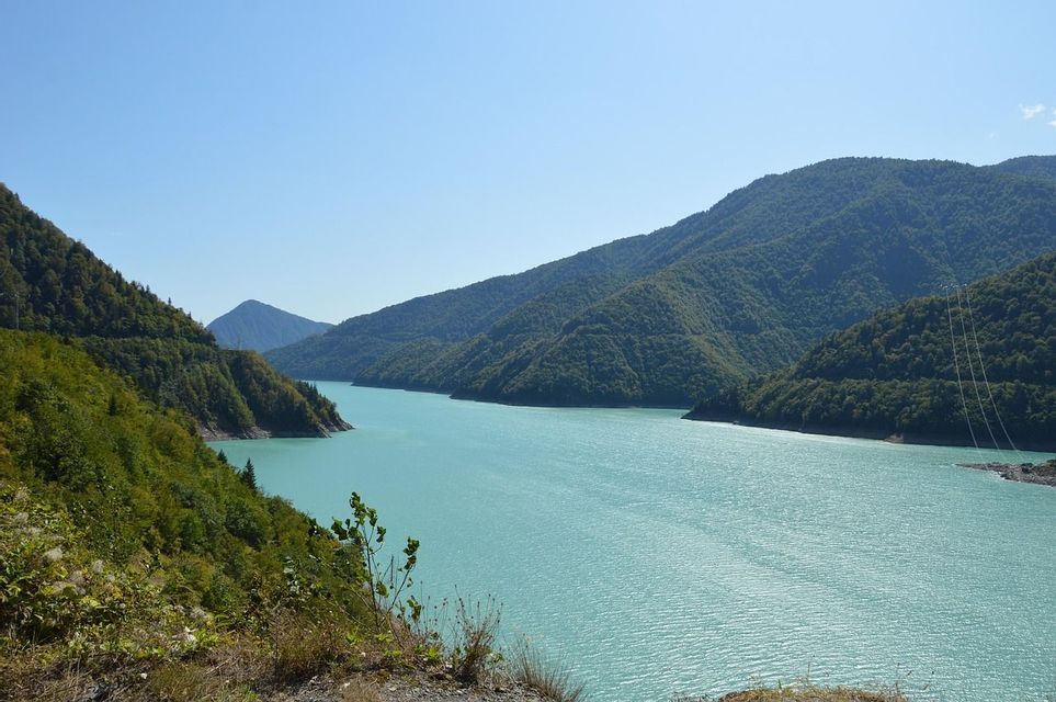 A wide turquoise river winds through a valley between steep, tree-covered mountains under a clear blue sky.