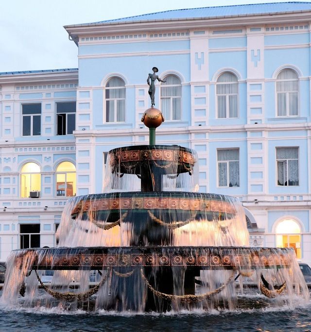An ornate, three-tiered fountain with a small statue on top, with water flowing in front of a large, light-blue building.