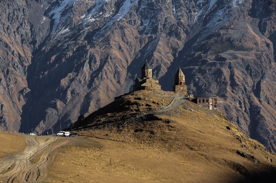 A stone church and bell tower stand on a grassy hill before a rugged, snow-capped mountain range.