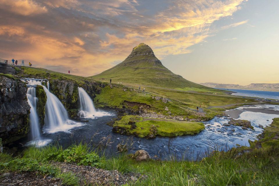 Wasserfälle stürzen eine üppig grüne Landschaft vor einem kegelförmigen Berg unter einem farbenprächtigen Sonnenuntergangshimmel hinab.