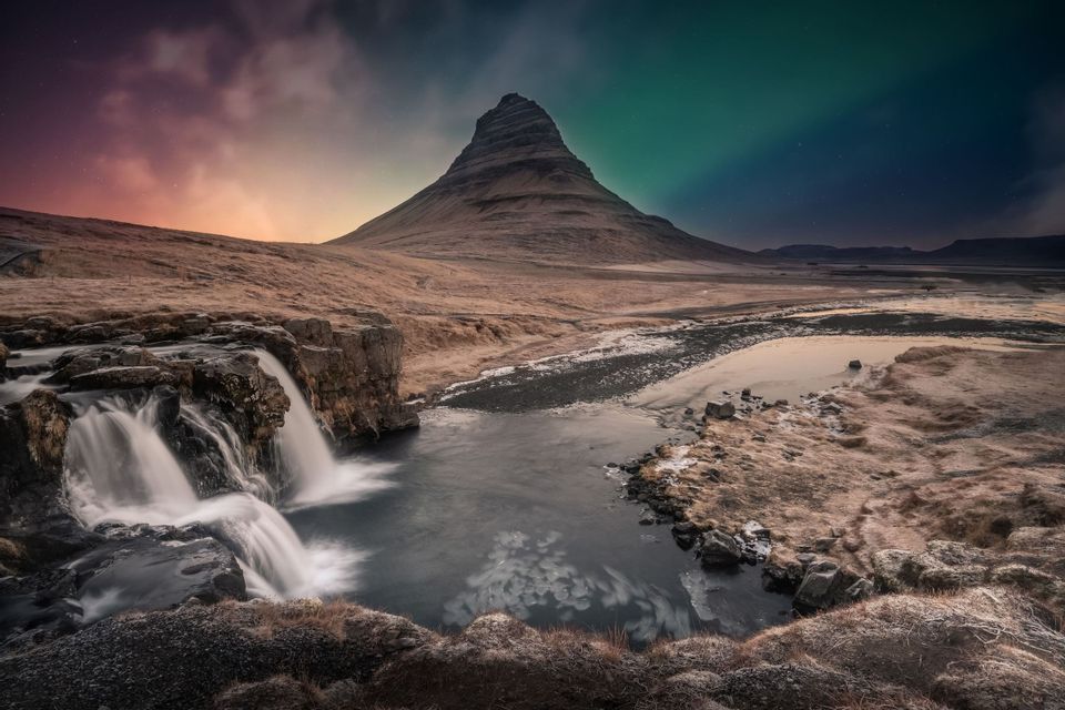 Ein kaskadierender Wasserfall in einem felsigen Tal mit einem großen Berg unter einem Nachthimmel mit Sternen und Nordlichtern.
