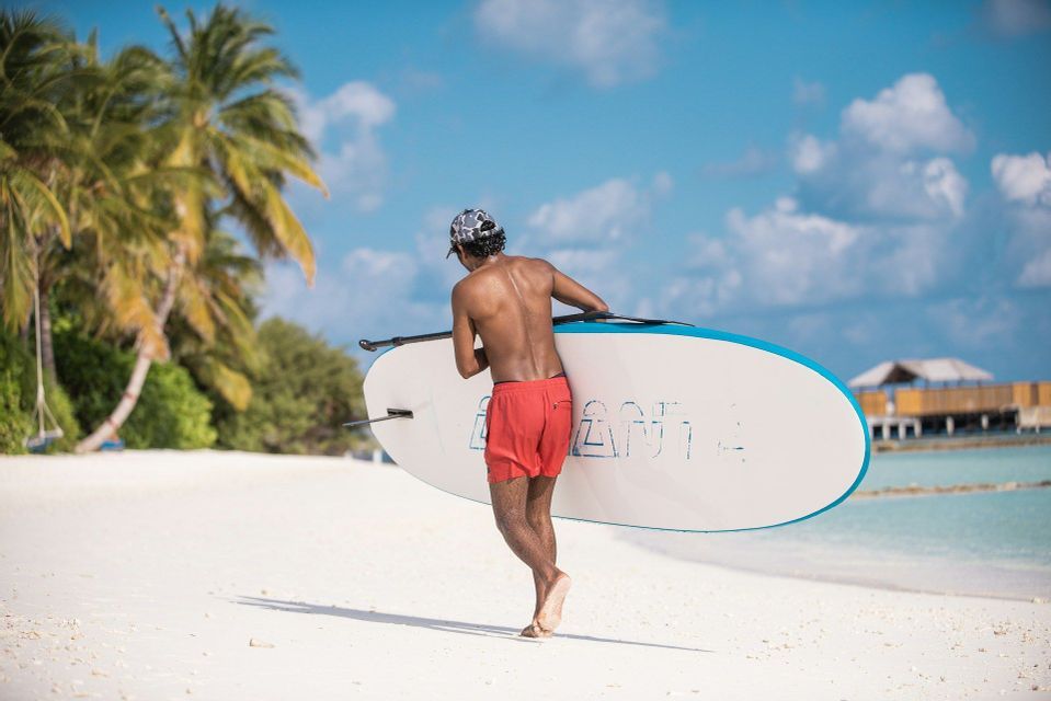Un hombre de espaldas, en bañador rojo, lleva una tabla de paddle surf mientras camina por una playa de arena blanca junto a agua turquesa y palmeras.