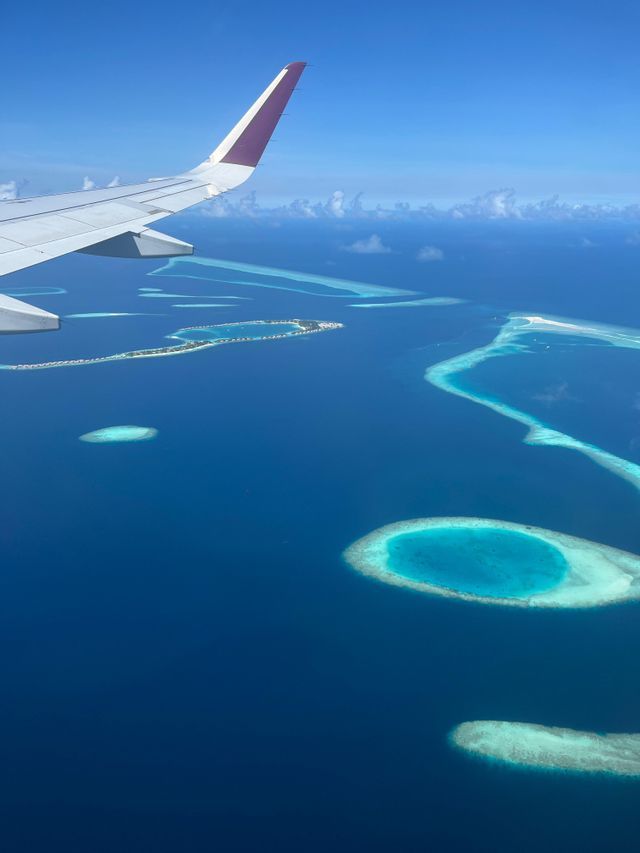 Una vista aérea desde la ventanilla de un avión, mostrando el ala sobre un océano azul profundo con varios atolones turquesa y bancos de arena.