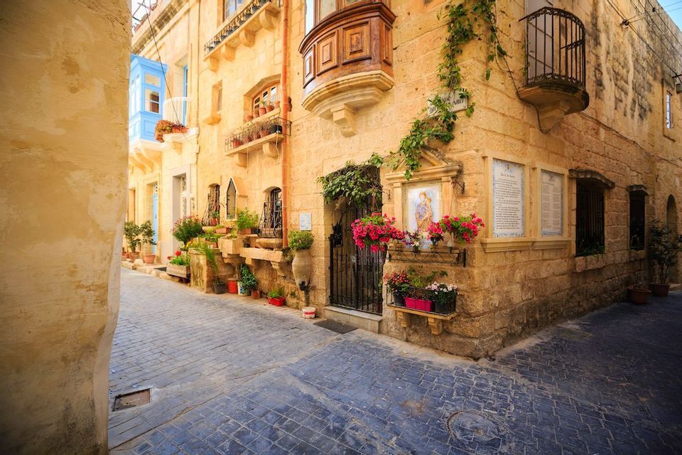A narrow cobblestone street winds past traditional stone buildings with enclosed wooden balconies and potted flowers.