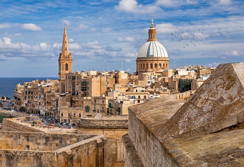 Una vista panoramica di una città costiera storica con una grande cupola e una guglia sotto un cielo parzialmente nuvoloso.