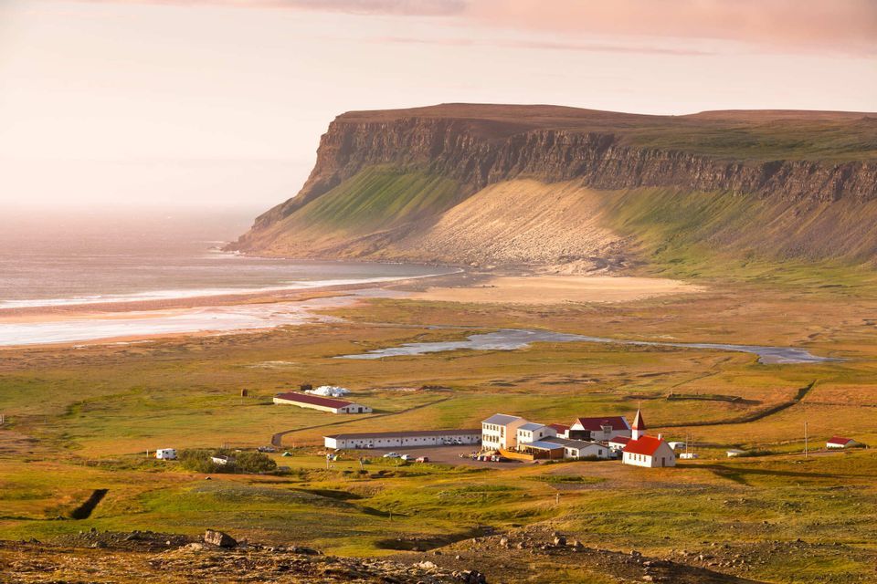 Ein kleines Küstendorf mit einer Kirche liegt auf einer grünen Ebene zwischen einem breiten Strand und einer großen, steilen Klippe.