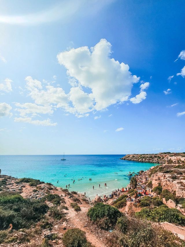 Una vista dall'alto di una spiaggia affollata in una cala rocciosa con acqua cristallina turchese sotto un cielo azzurro brillante.