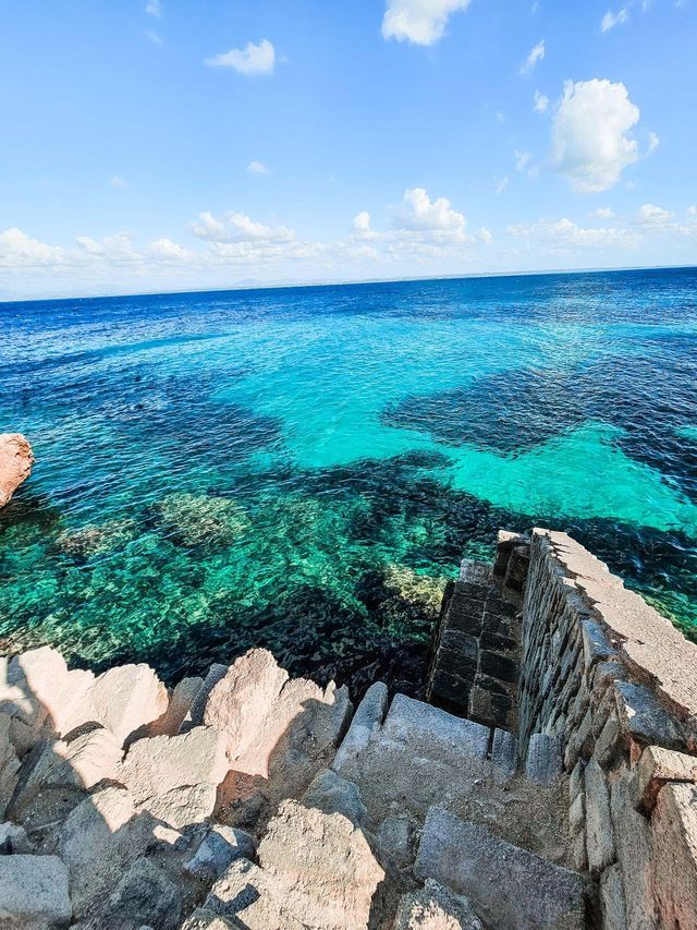 Gradini di pietra su una costa rocciosa scendono verso un mare limpido turchese sotto un cielo azzurro brillante con nuvole rade.